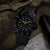 Herrenuhr am Handgelenk mit schwarzem Armband und Leuchtziffern auf dunklem Zifferblatt.