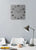Quadratische Wanduhr an der Wand mit grauem Ziffernblatt und silbernen Zahlen.